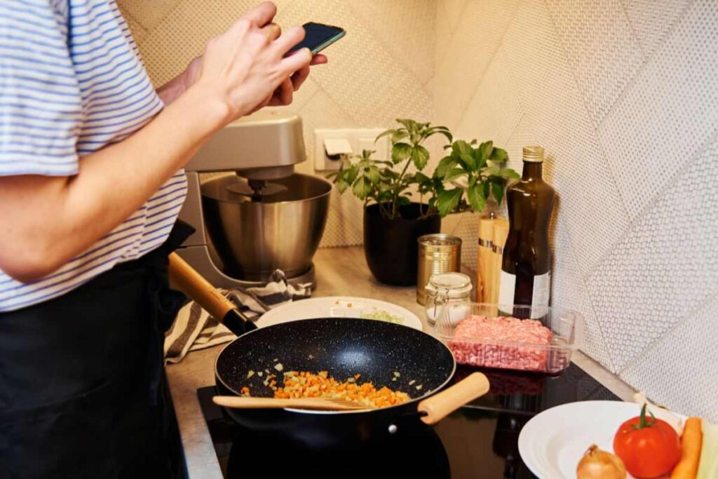 donna ai fornelli mentre cucina e tiene smartphone in mano